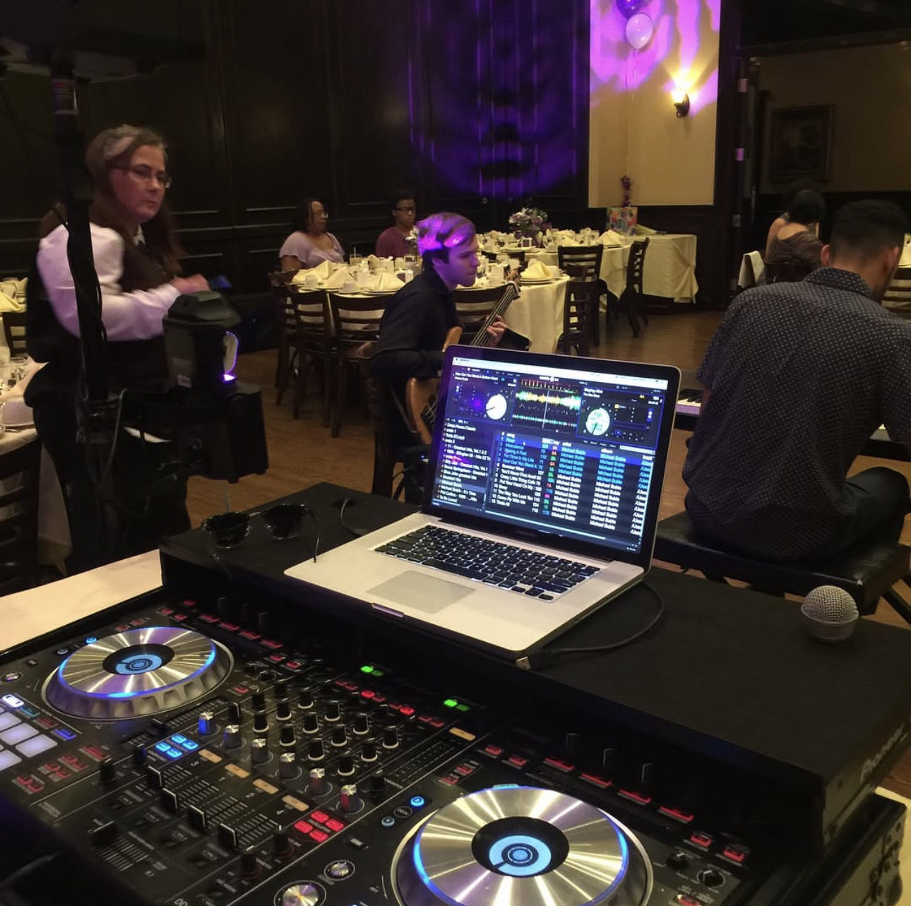A laptop and controller set up at the front of a banquet room lit purple, guests seated at tables behind.