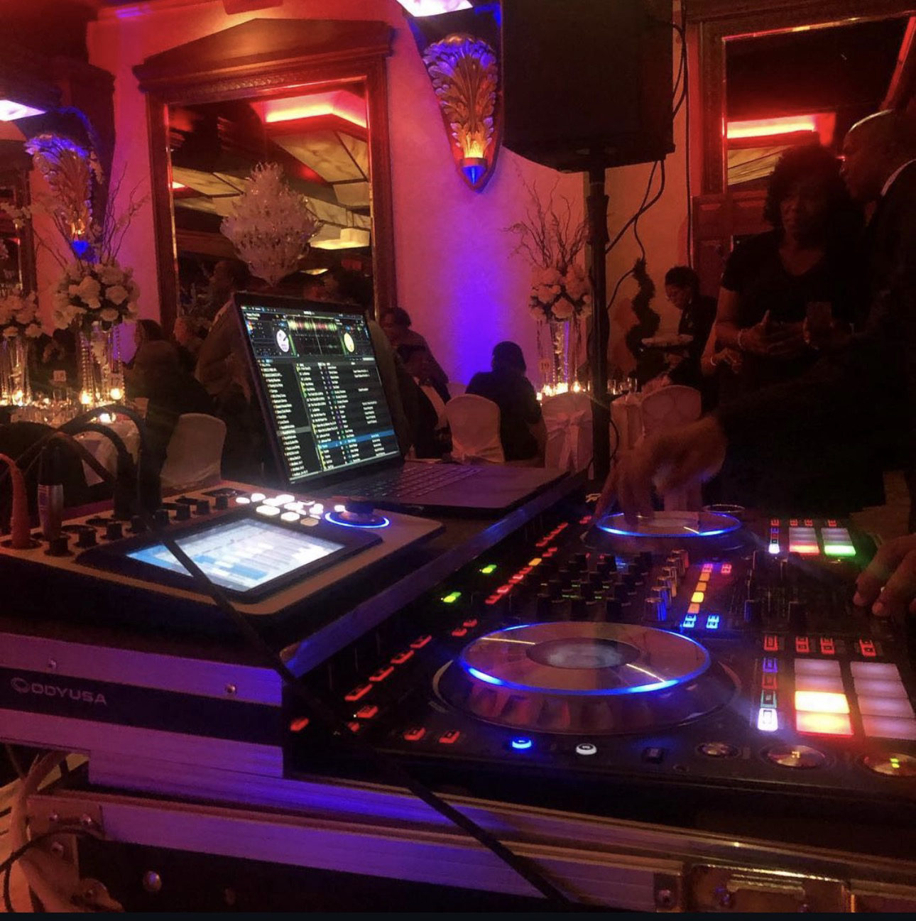 A DJ controller lit blue at the edge of a wedding reception, the room washed in warm red and purple uplighting.